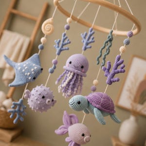 May include: A nursery mobile featuring a circular wooden frame with hanging crochet sea creatures including a blue stingray, a purple pufferfish, a purple jellyfish, a purple and blue sea turtle, and a purple fish, accented with coral and shell shapes.