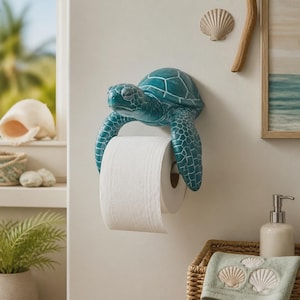 Op de afbeelding: Een aan de muur gemonteerde toiletpapierhouder in de vorm van een zeeschildpad met een blauwgroen schild en een getextureerde huid. De voorste flippers van de schildpad hangen naar beneden en ondersteunen een standaard rol wit toiletpapier.