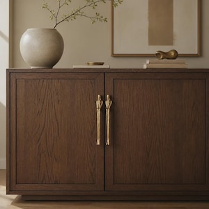 May include: A dark brown wooden sideboard featuring two cabinet doors with vertical brass handles shaped like elongated human figures. The cabinet has a rectangular frame design on each door and a smooth, natural wood grain finish throughout the surface.