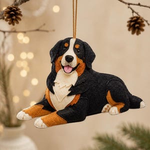 Könnte beinhalten: Ein handgefertigter Filz-Anhänger in Form eines Berner Sennenhundes mit dem typischen schwarz-braun-weißen Fell. Der Hund ist in liegender Position dargestellt, mit fröhlichem Gesichtsausdruck, rosa Zunge und einer goldenen Aufhängeschlaufe.