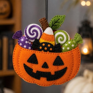 Puede incluir: Un adorno colgante de fieltro con forma de cubo de calabaza naranja lleno de dulces variados, incluyendo una piruleta de espiral morada, una piruleta de espiral verde, un caramelo de maíz y dos caramelos envueltos con lunares morados y verdes.
