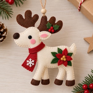 Könnte beinhalten: Ein Rentier-Anhänger aus Filz in Creme mit dunkelbraunem Geweih und Hufen. Das Rentier trägt einen roten Schal mit weißer Schneeflocken-Applikation und eine rote Weihnachtsstern-Blüte mit grünen Blättern an der Seite. Ein kleiner Zweig Stechpalme ziert das Geweih.