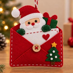 May include: A red felt envelope ornament featuring a Santa Claus figure with a white beard and red hat, holding a green gift. The envelope is decorated with a small red heart, a wooden button closure, and a green Christmas tree with a yellow star.
