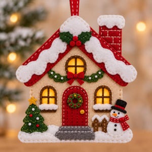Peut inclure: Une décoration de Noël en feutrine en forme de maison avec un toit rouge couvert de neige blanche, une porte rouge avec une couronne, deux fenêtres lumineuses, une cheminée, un sapin vert avec une étoile jaune et un bonhomme de neige.