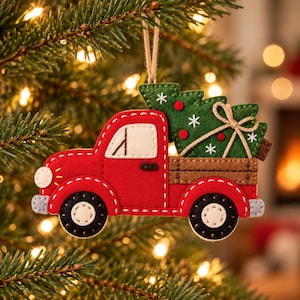 Puede incluir: Un adorno de fieltro para árbol de Navidad con forma de camioneta roja que lleva un árbol de Navidad verde en su caja marrón. La camioneta tiene ruedas blancas, una ventana blanca y un lazo de cuerda de yute para colgarla.
