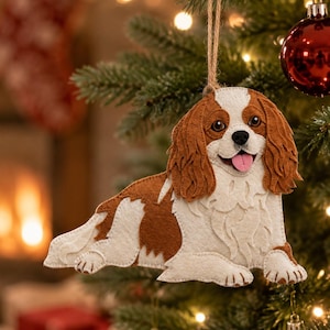 May include: A felt Cavalier King Charles Spaniel ornament featuring brown and white layered fabric details, a pink tongue, and a brown twine loop for hanging. The dog is depicted in a lying position with long, textured ears and dark eyes.