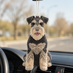 Puede incluir: Un adorno colgante de fieltro con forma de perro Schnauzer con detalles de pelaje gris carbón oscuro y beige claro. El perro tiene un borde cosido, ojos negros, una lengua rosa y está suspendido por un cordel blanco fino.
