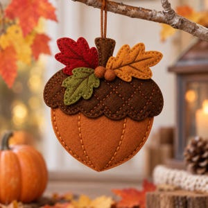 May include: A felt acorn ornament featuring a dark brown textured cap and a light orange base with stitched vertical lines. The top is decorated with two small felt balls and three felt leaves in red, green, and golden yellow colours.