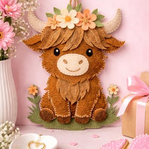 May include: A brown felt Highland cow wall hanging featuring a cream-colored muzzle, black eyes, and a crown of felt flowers in shades of peach, cream, and green. The cow sits on a green felt patch with small flowers and hangs from a brown cord.