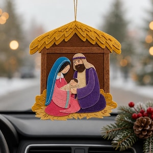 May include: A felt hanging ornament depicting a nativity scene with a yellow thatched roof, brown stable walls, and figures of a person in a blue head covering and pink dress, a person in a purple robe, and a small infant wrapped in white cloth.