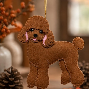 May include: A brown felt poodle ornament with a gold hanging cord. The dog figure features stitched edges, black eyes, a black nose, and light pink inner ears. The poodle is depicted in a standing position with a curly tail and a small tongue.