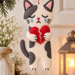 Peut inclure: Une décoration en feutrine en forme de chat au pelage crème et gris, avec des yeux fermés, des larmes bleues et un nez rose. Le chat tient un cœur brisé rouge entre ses pattes et est suspendu par une ficelle torsadée rouge et blanche.