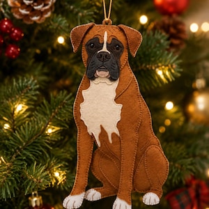May include: A felt Boxer dog ornament featuring a brown coat with a white chest patch, white paws, and a black muzzle. The dog is depicted in a sitting position with a gold hanging string attached to the top, showcasing detailed stitching.
