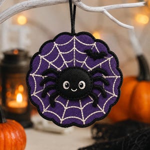 May include: A circular purple felt ornament featuring a white stitched spiderweb pattern and a central black spider with white eyes and a small smile. A small black bat is stitched near the top edge, and a black ribbon loop is attached for hanging.