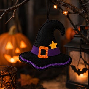 May include: A black felt witch hat ornament featuring a purple band, an orange square buckle, and a small yellow star accent. The hat is stitched with purple thread along the brim and hangs from a thin black cord for display purposes.