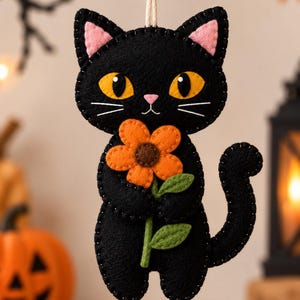 May include: A black felt cat ornament featuring large yellow eyes, pink inner ears, and a small pink nose. The cat holds an orange flower with a brown centre and green leaves. The edges are stitched with black thread, and a string with a wooden bead hangs from the top.