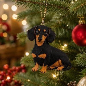 Peut inclure: Une décoration en forme de teckel noir et feu fabriquée en feutrine avec des coutures visibles sur les bords. Le chien est en position assise et comporte une chaînette à billes en métal doré fixée au sommet pour l'accrocher aux branches.