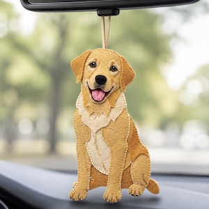 May include: A felt hanging ornament shaped like a golden retriever dog with yellow and tan layered fabric pieces, a black nose, and a pink tongue, suspended by a thin tan cord from a black rearview mirror mount in a vehicle interior.