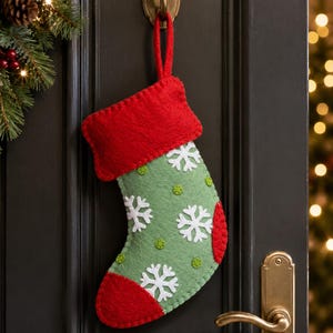 May include: A felt Christmas stocking featuring a sage green body with white snowflake appliqués and small lime green circular accents. The stocking has a bright red cuff, a red toe, a red heel, and a matching red hanging loop made of felt.