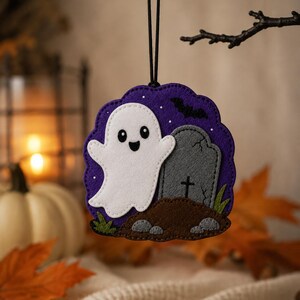 May include: A felt hanging ornament featuring a white ghost next to a gray tombstone on a brown mound of dirt, set against a purple background with a black bat silhouette, suspended by a thin black cord for display as a seasonal decoration.