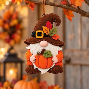 Può includere: Un ornamento in feltro a forma di gnomo con un cappello marrone, una fascia nera, una fibbia gialla e foglie in tessuto colorate. Lo gnomo ha una barba bianca, mani color pesca e tiene in mano una zucca arancione brillante con foglia.