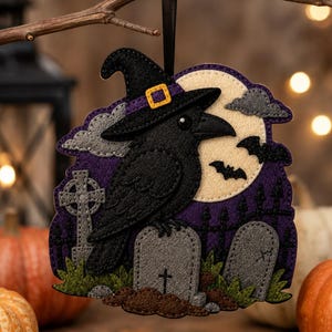 May include: A felt hanging ornament featuring a black raven wearing a witch hat, perched on a gray tombstone. The background displays a full moon, purple sky, gray clouds, bats, a cross, and a black fence, all crafted from layered felt pieces.