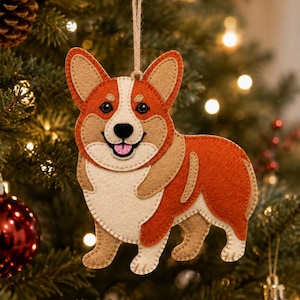 May include: A felt Corgi ornament featuring a tan and orange dog with a white chest and face markings. The ornament has visible stitching along the edges and is suspended by a thin, light brown twine loop for hanging on a tree branch.