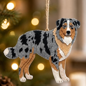 May include: A felt Australian Shepherd dog ornament featuring a blue merle coat with black patches, tan accents on the legs and face, and a white chest. The dog is depicted in a standing profile and hangs from a thin, light brown jute twine loop.