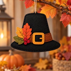 May include: A black felt pilgrim hat ornament featuring a brown band with a yellow buckle, decorated with three felt leaves in orange, yellow, and olive green, and three small red berries. The ornament hangs from a thin, light brown twine loop.