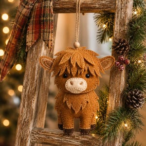 May include: A brown felt Highland cow ornament featuring a cream-coloured muzzle, dark brown hooves, and black bead eyes. The ornament is suspended by a thin jute twine string with a small wooden bead at the top, showcasing detailed stitching.