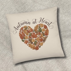 Könnte beinhalten: Ein quadratisches Dekokissen mit cremefarbenem Bezug, das ein herzförmiges Arrangement aus verschiedenen Herbstblättern in Orange-, Gelb-, Braun- und Grüntönen zeigt. Der Text "Autumn at Heart" ist in schwarzer Schreibschrift über dem Herz gedruckt.