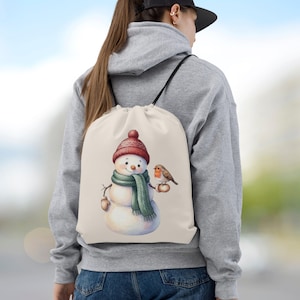 Könnte beinhalten: Ein cremefarbener Kordelzugrucksack mit einer grafischen Illustration eines Schneemanns, der eine rote Strickmütze und einen grünen Schal trägt, wobei ein kleines braunes Rotkehlchen auf einem seiner Stockarme sitzt. Schwarze Kordelträger.