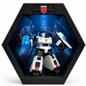 May include: A Megatron action figure displayed inside a black hexagonal frame. The figure is white, black, and grey with a red Decepticon emblem on its chest and a blue translucent cannon attached to its arm. The frame features a matching red emblem.