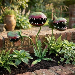 May include: A metal garden sculpture featuring three stylized carnivorous plants with open, toothy mouths. The plant heads are painted in shades of green and reddish-purple, supported by thin green stems and jagged, pointed leaves at the base.