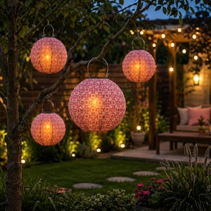 May include: Four spherical hanging lanterns with a pink and white geometric pattern, each featuring a metal handle and an internal warm light source, suspended from the branches of a tree in an outdoor garden setting at twilight.