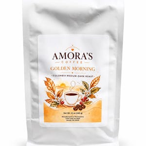 May include: A white coffee bag featuring a label for Amora's Coffee Golden Morning Colombia medium-dark roast. The label displays a cup of coffee, coffee beans, and autumn-colored leaves. Net weight is 12 oz (340 g). Manufactured in Dacula, GA.