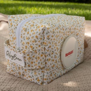 May include: A rectangular fabric pouch featuring a yellow and green floral pattern on a white background. The pouch has a white zipper, a side handle with a white tag labeled RileyBean, and a circular white plastic dispenser lid labeled HUGGIES.