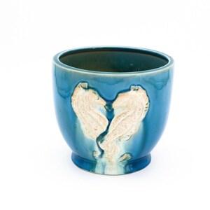 May include: A deep teal ceramic planter featuring a raised, textured cream-colored relief of two seahorses facing each other to form a heart shape. The vessel has a smooth, glossy finish with a rounded body and a small, circular pedestal base.