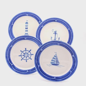 Puede incluir: Un juego de cuatro platos redondos de cerámica con centro color crema y un borde azul ancho con un patrón decorativo de cuerda. Cada plato muestra una ilustración náutica azul distinta: un faro, un ancla, un timón y un velero.
