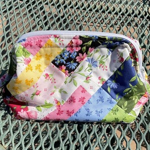 May include: A quilted zippered pouch featuring a diagonal patchwork design of various floral fabrics in shades of yellow, pink, blue, green, and white. The pouch has a white zipper closure along the top edge and a soft, padded construction.