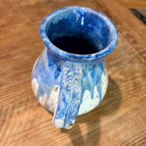 May include: A ceramic pitcher featuring a glossy blue glaze that drips down over a speckled cream-colored base. The handle displays a textured, leaf-like pattern carved into the surface, and the interior of the vessel is finished in a deep blue.