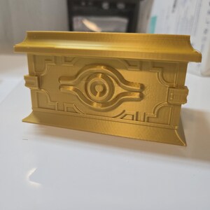 Könnte beinhalten: Eine goldfarbene rechteckige Box mit 3D-Druck-Finish. Die Vorderseite zeigt ein erhabenes, symmetrisches geometrisches Design, das um ein kreisförmiges Motiv mit horizontalen Erweiterungen zentriert ist und an ein antikes Artefakt erinnert.