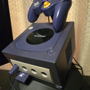 May include: A purple Nintendo GameCube video game console with a matching purple controller resting on top. The console features a black circular disc cover with the GameCube logo, a front-facing memory card slot with a purple card, and controller ports.