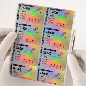 May include: A roll of rectangular adhesive labels with a holographic finish. Each label features black text reading TB-500, 10mg, Purity ≥ 99%, Exp: 08/26, Lot: 24A15, and For Research Use Only, along with a small DNA strand graphic and icons.