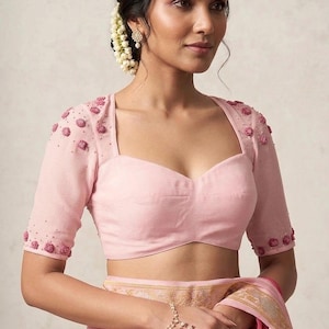 May include: A light pink blouse featuring a sweetheart neckline and short sleeves with sheer mesh overlays. The sleeves are embellished with small pink floral appliqués and tiny beads. The blouse is paired with a matching pink fabric draped garment.