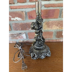 May include: A metallic bronze-toned lamp base featuring a sculpted cherub figure wrapped around the central column. The base has an ornate, flared design with intricate detailing, a cylindrical top socket, and a brown electrical cord with a plug.
