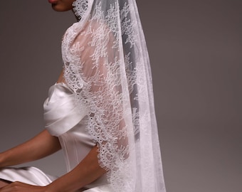 May include: A long white bridal veil featuring intricate floral lace detailing along the edges. The sheer fabric drapes over the head and shoulders, showcasing a delicate scalloped lace trim that frames the face and extends down the length.