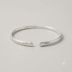 May include: A silver-colored open cuff bracelet featuring a hammered, textured surface throughout the band. The bracelet has two tapered, pointed ends that face each other with a small gap in between, creating a simple, circular silhouette.