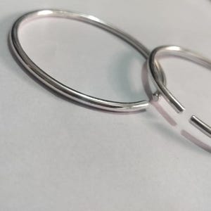 May include: Two circular silver-toned hoop earrings with a smooth, polished finish. Each hoop features a simple, open-ended design with a thin wire construction, reflecting light across the metallic surface of the jewelry pieces.