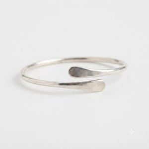 May include: A silver-toned open cuff bracelet with a smooth, polished finish. The bracelet features a circular band that does not fully close, ending in two slightly flattened, rounded terminals that face each other with a small gap.